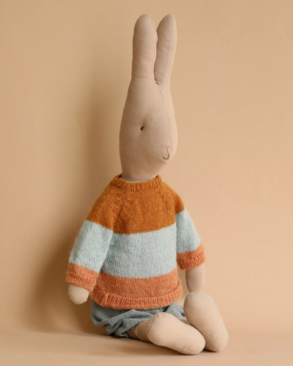A Maileg Rabbit size 5, Classic - Sweater and Shorts sits against a beige background. The rabbit is wearing a knitted sweater with orange and blue horizontal stripes. Its legs are clad in light-colored cotton fabric, likely shorts or a skirt. The serene and simple design of the rabbit's wardrobe adds to its charm.