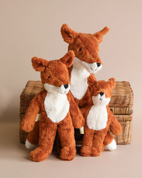 Three Senger Naturwelt Stuffed Animal - Fox toys of varying sizes posed on a wicker basket against a light beige background. The toys have brown and white fur with blue eyes.