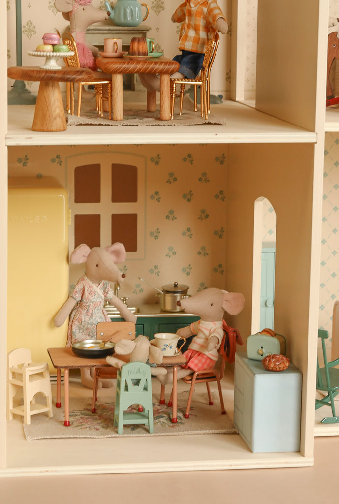 Two toy mice, dressed in clothes, sit at a kitchen table inside the Maileg Fully Furnished Dollhouse Set + Surprise Gift, featuring mini furniture like a yellow fridge, stove, cabinets, and playful household items for a cozy scene.