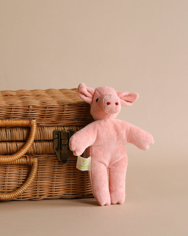 A Senger Naturwelt Stuffed Animal - Baby Pig standing beside a closed wicker basket on a neutral beige background. The pig toy features a soft texture and smiling facial expression, crafted from organically grown cotton.