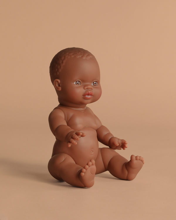 The Minikane Doll (13") - Zélie sits upright on a solid beige background, showcasing brown skin, short textured hair, and detailed facial features. Anatomically correct and made in Spain. Clothing sold separately.