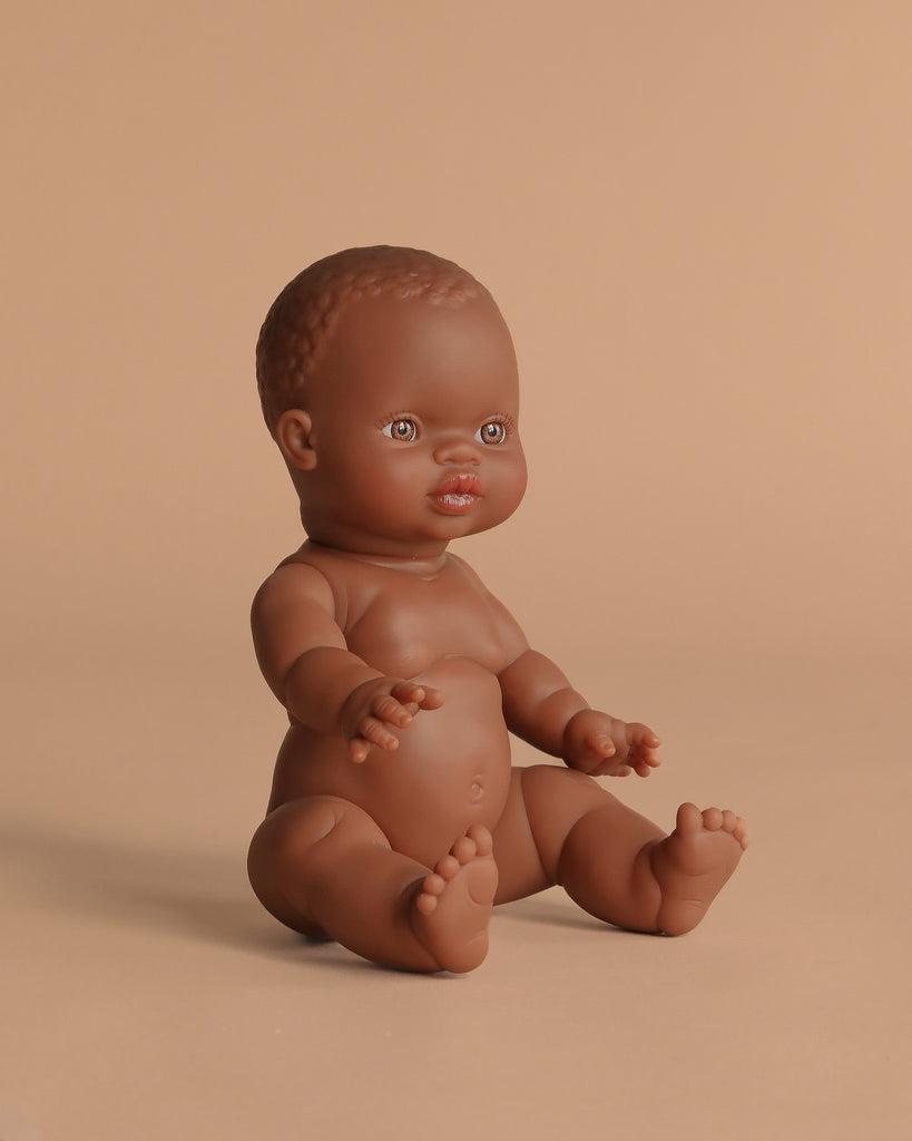 The Minikane Doll (13") - Zélie sits upright on a solid beige background, showcasing brown skin, short textured hair, and detailed facial features. Anatomically correct and made in Spain. Clothing sold separately.