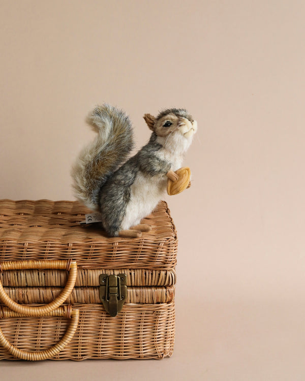A Squirrel With A Nut Stuffed Animal toy with a fluffy tail, meticulously hand-sewn, sits atop a wicker picnic basket against a soft beige background, portraying a cute and whimsical scene.