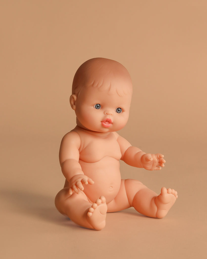 The Minikane Vintage Baby Doll (13") - Lily, with light skin, blue eyes, and molded hair, sits nude on a light beige background with its legs spread and arms extended forward. Clothing sold separately. Made in Spain.