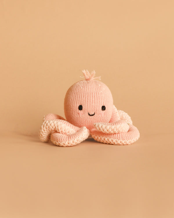 The Cuddle + Kind Baby Octopus (Soft Coral Pink), a hand-knit heirloom toy made from Peruvian cotton yarn with a smiling face, rests on a plain beige background.