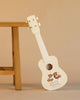 The Konges Sloejd Wooden Ukulele - Teddy Bear, a small white wooden ukulele featuring a charming teddy bear design, leans against a wooden table on a beige background, making it an adorable FSC-certified kids’ musical instrument.