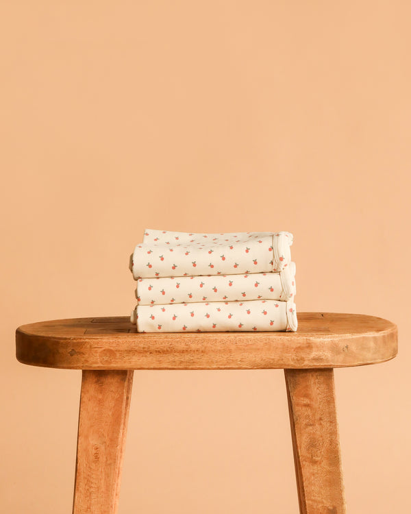 The Organic Baby Blanket – Little Peach, made from GOTS-certified organic cotton with a cream base and small red floral pattern, is neatly folded on a wooden bench against a peach background for a gentle, chemical-free touch.