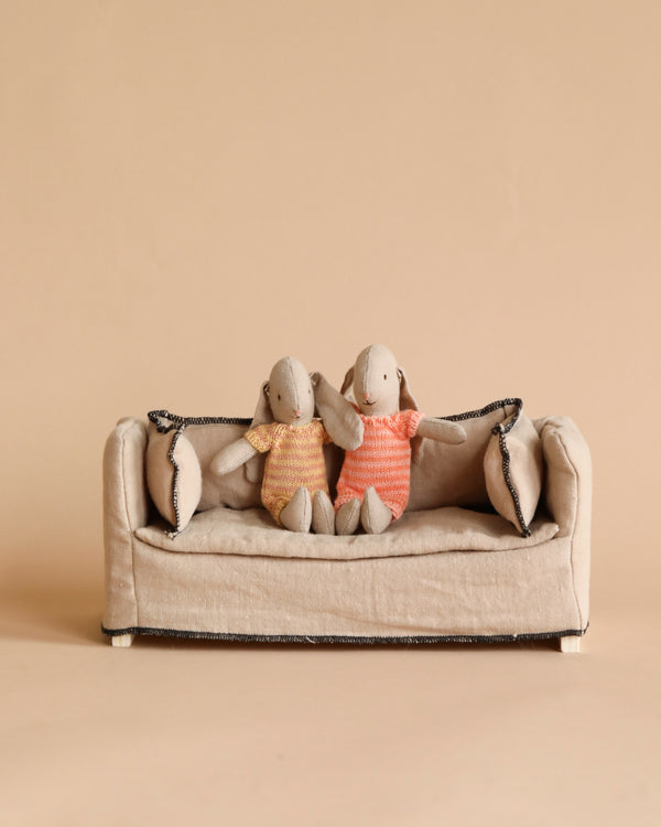 Two Maileg Micro Bunnies in striped suits sitting on a miniature beige sofa against a plain beige background.