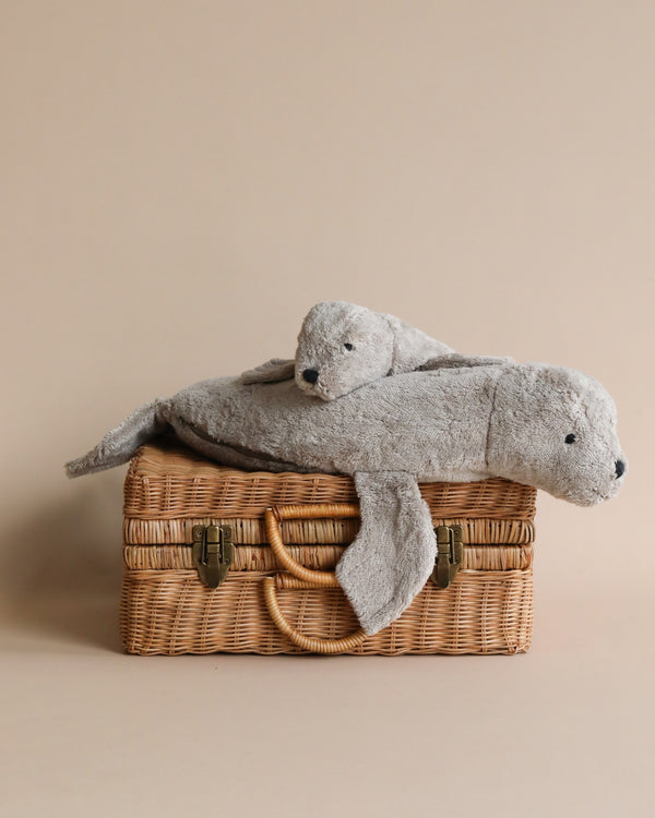 A Senger Naturwelt Cuddly Animal - Gray Seal, lying on top of a small woven wicker basket, set against a plain beige background. The seal is gray and appears soft, with a relaxed