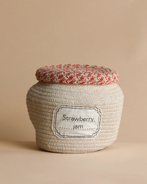 A fabric-wrapped jar labeled "Strawberry Jam Jar Basket" with a textured beige body and a red and white speckled fabric lid against a soft beige background.