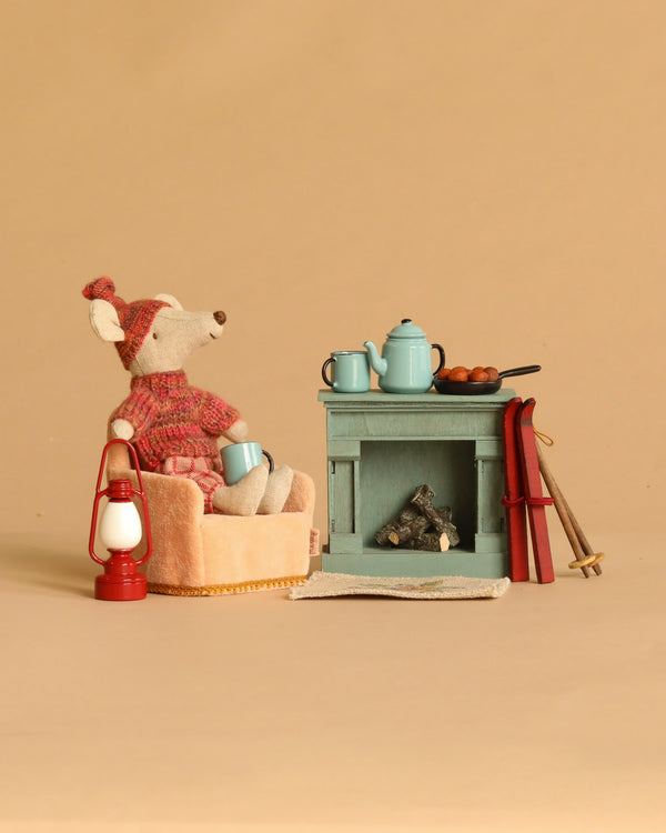 A Maileg Winter Chalet Set features a Winter Ski Mouse in a pink chair holding a blue mug beside a cozy Maileg Fireplace with a blue teapot, pan, and cups. Nearby are a Maileg Lantern, rug, red skis, and ski poles on a beige background.