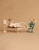 The Maileg Bedroom Set with Mum & Dad is showcased in a minimal room: one mouse dressed in pajamas sits up in the cozy bed complete with a quilt, while the other, wearing a robe and glasses, relaxes on the rocking chair beside a softly glowing lamp.