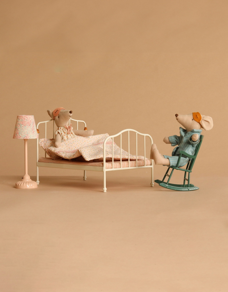 The Maileg Bedroom Set with Mum & Dad is showcased in a minimal room: one mouse dressed in pajamas sits up in the cozy bed complete with a quilt, while the other, wearing a robe and glasses, relaxes on the rocking chair beside a softly glowing lamp.
