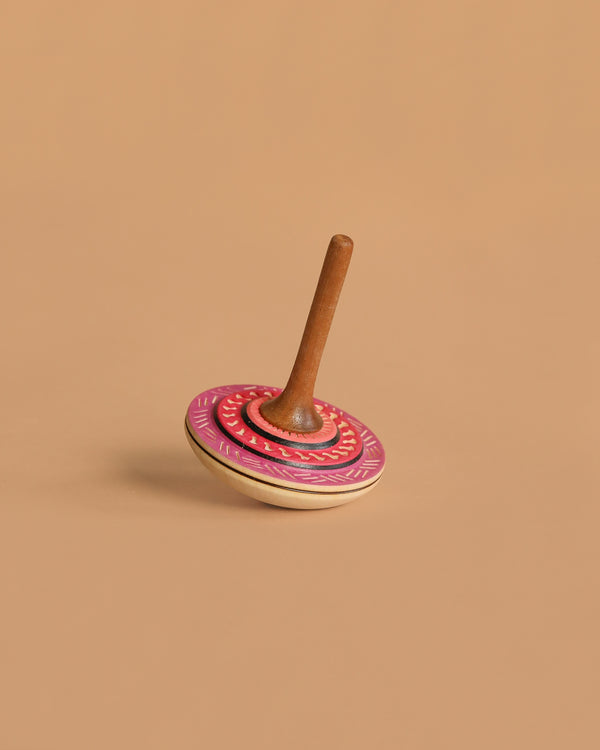 The Mader Bonbon Spinning Top - Pink features an embossed design and a decorated pink and red circular base with a brown handle, photographed standing upright on a plain beige surface.