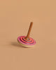 The Mader Bonbon Spinning Top - Pink features an embossed design and a decorated pink and red circular base with a brown handle, photographed standing upright on a plain beige surface.