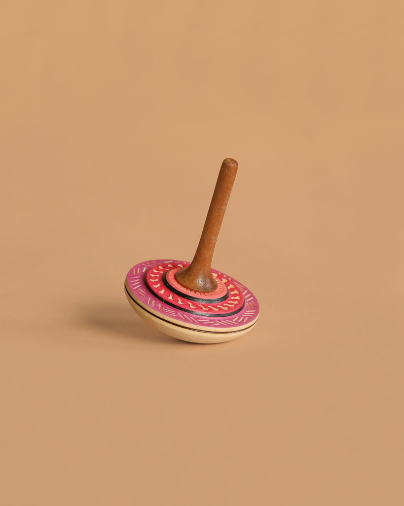 The Mader Bonbon Spinning Top - Pink features an embossed design and a decorated pink and red circular base with a brown handle, photographed standing upright on a plain beige surface.
