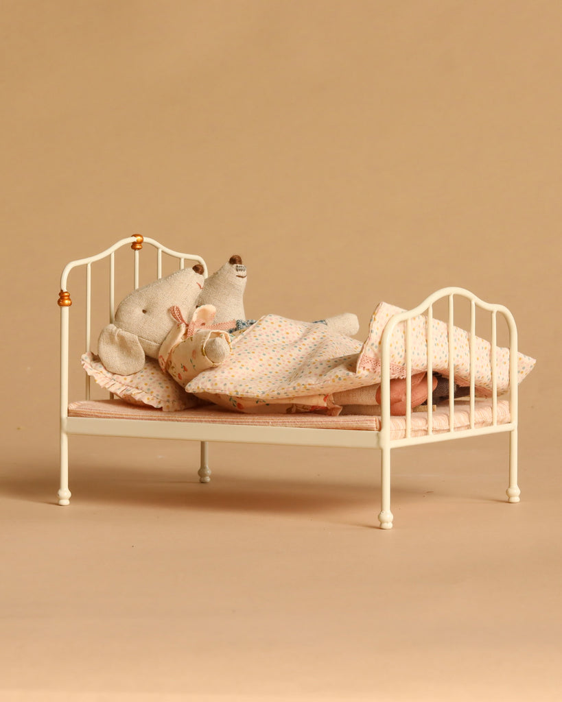Two Maileg mice, Grandma and Grandpa, from the Maileg Bedroom Set with Grandma & Grandpa, are dressed in pajamas and lying together under a polka-dot blanket on a vintage bed with pink bedding, set against a plain beige background.