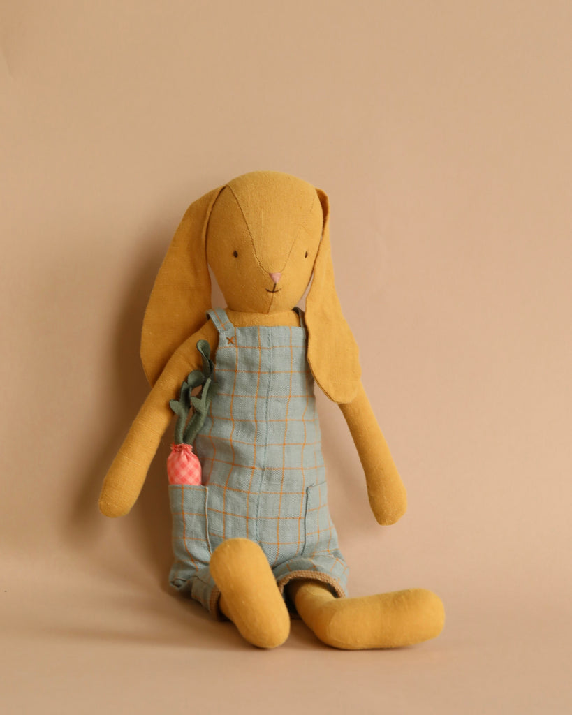 A Maileg Bunny Size 3 plush bunny toy with droopy ears, wearing checkered overalls from its bunny wardrobe and holding a small pink crochet detail, sitting against a plain beige background.