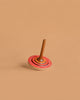 The Mader Bonbon Spinning Top - Orange features a wooden design with an orange, embossed pattern disc and a brown handle, shown against a plain beige background.