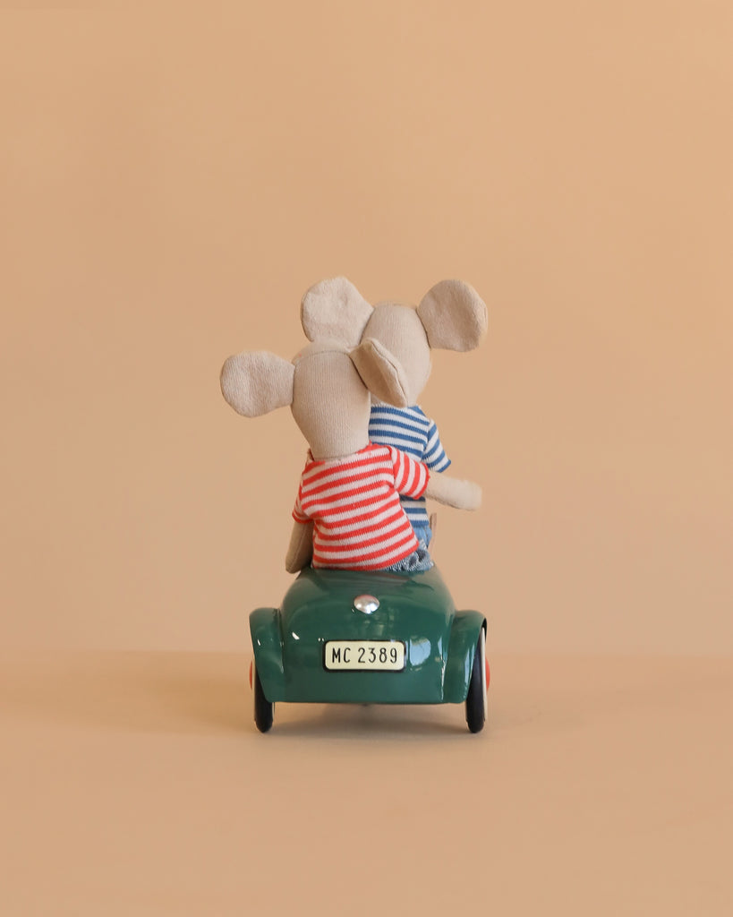 The Maileg Big Brother and Sister Adventure Set features two plush mice in striped shirts riding together in a small green car, seen from behind against a plain beige background.