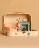 A Maileg Princess Mouse in a pink dress stands in a decorated scene featuring the Maileg On The Go Suitcase Set, complete with Maileg Dollhouse Furniture such as a pink chair, green fireplace, and a clock on the mantel, all set against a beige background.