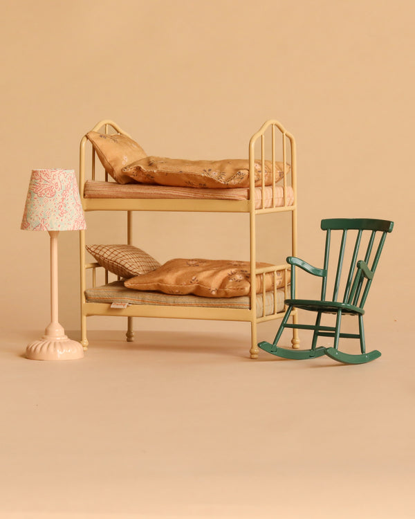 A miniature scene featuring the Maileg Bunk Bed Bedroom Set with a cream finish and patterned bedding, complemented by a pink floral Maileg Vintage Floor Lamp and a green Maileg Rocking Chair, all set against a plain beige background.