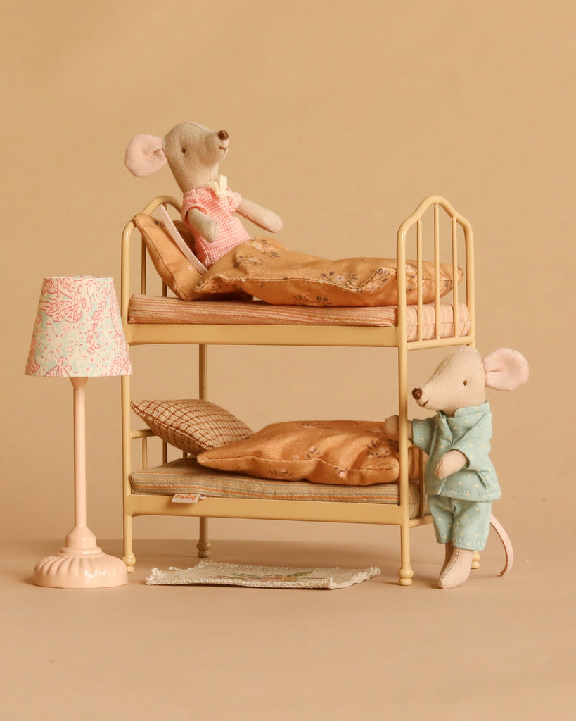 Two Maileg mice, Little Brother and Sister, dressed in pajamas, are posed with the Maileg Bunk Bed Bedroom Set. One mouse is lying on the top bunk while the other stands beside the lower bunk as if ready to climb in. A tiny lamp and a soft beige background complete the cozy bedtime scene.