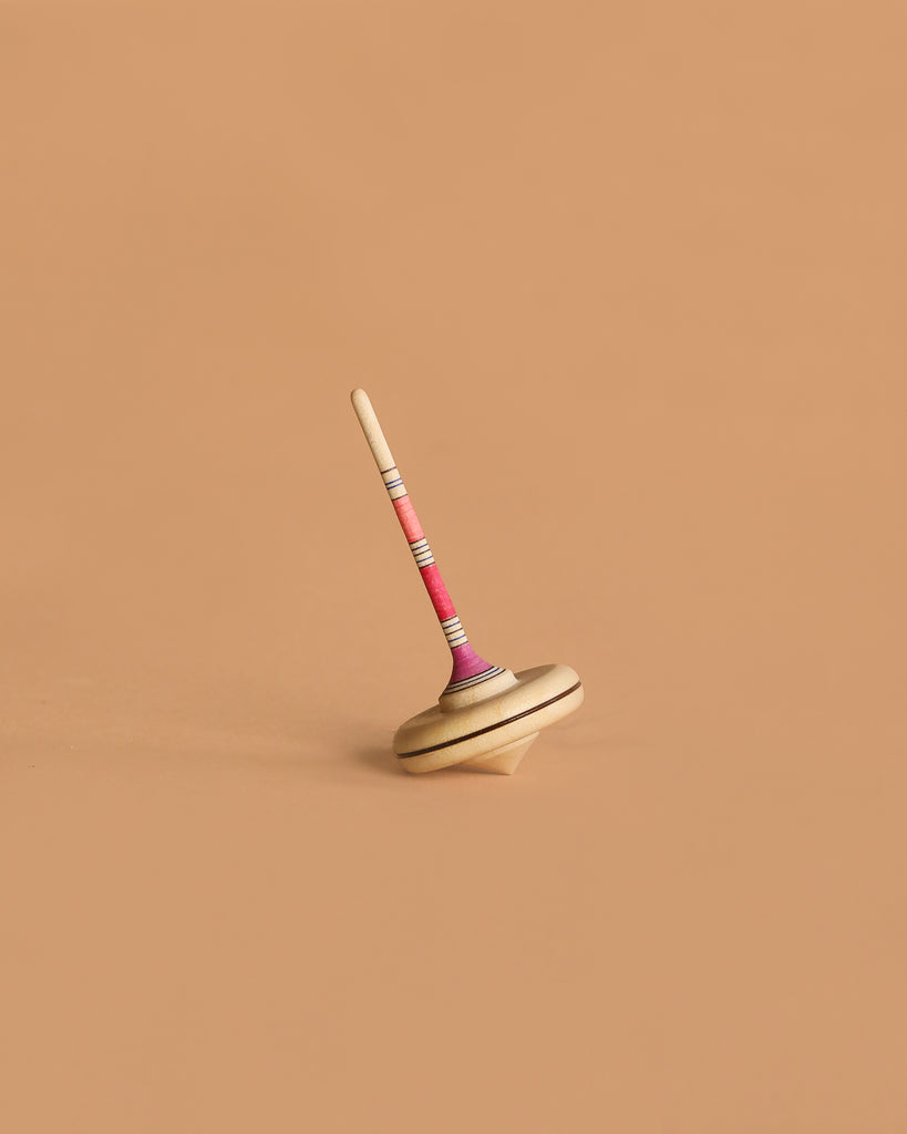 The Mader Spaghetti Spinning Top - Pink, crafted from maple wood, is balanced upright on a plain, light brown surface.
