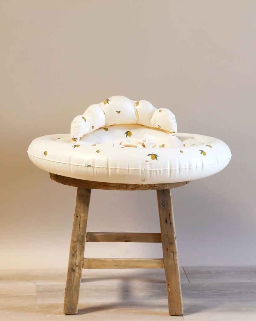 A round, durable Inflatable Baby Swim Ring - Lemon with a white and leafy pattern placed on an aged wooden stool against a neutral beige background.