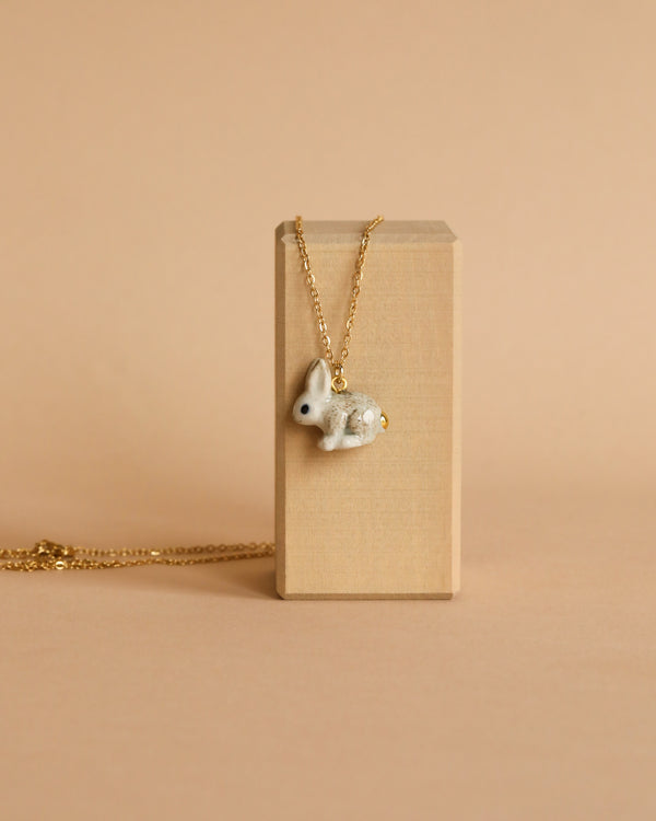 A Goldentail Rabbit Necklace with a small, detailed porcelain pendant shaped like a rabbit is draped over a beige, rectangular wooden block. The background and the block are both in a neutral, tan shade, which complements the 24k gold-plated chain and highlights the intricate rabbit pendant.