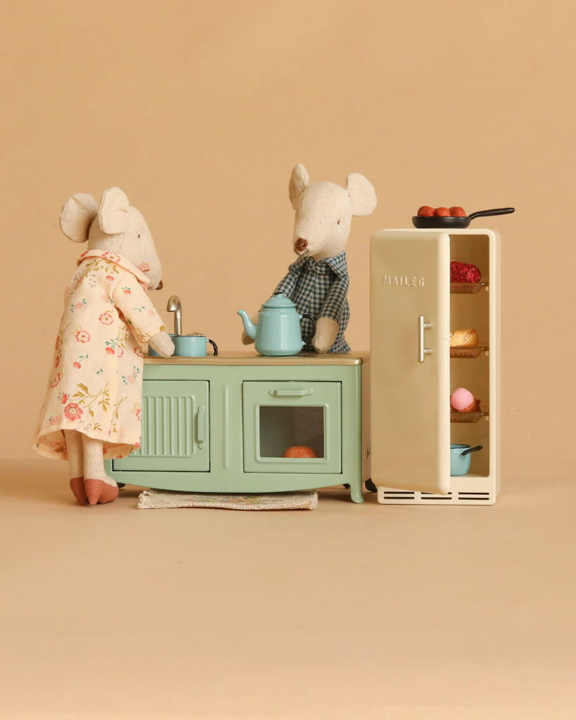 Grandma and Grandpa Maileg mice stand beside their Maileg Kitchen Set, which includes a metal fridge stocked with food and a stove with a pan. The scene is set against a solid beige background.