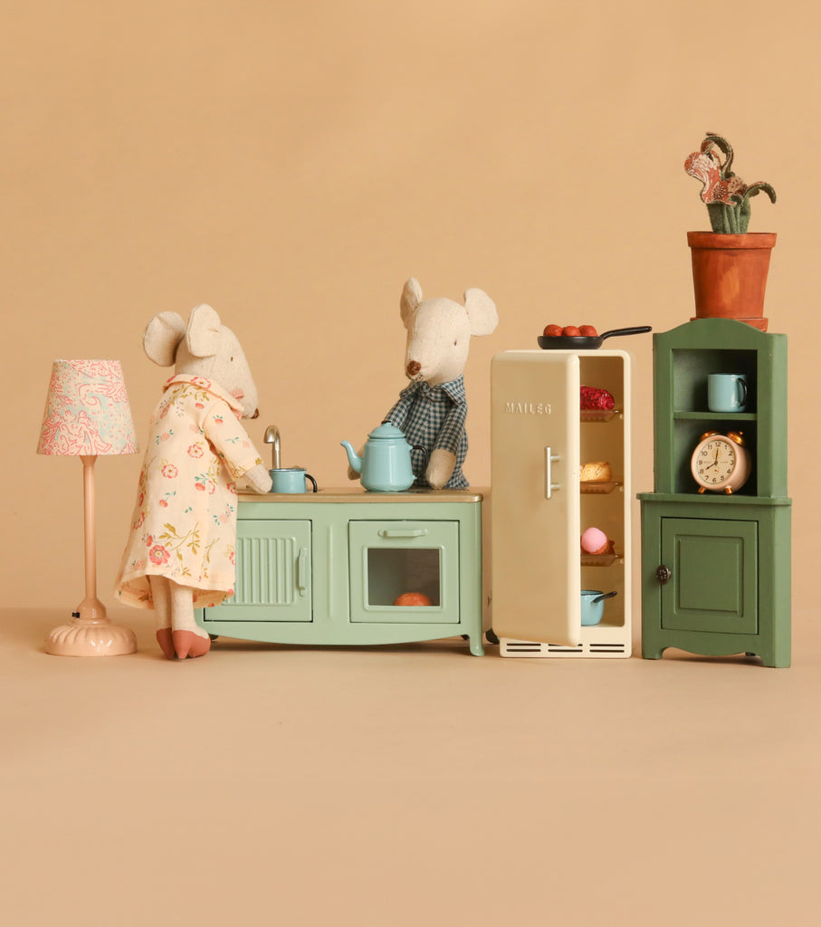 Grandma and Grandpa Maileg mice enjoy a cozy time at home in a delightful miniature kitchen, featuring the Maileg Kitchen Set with pastel stove, open fridge, cabinet, lamp, and potted plant—all set against a beige background.