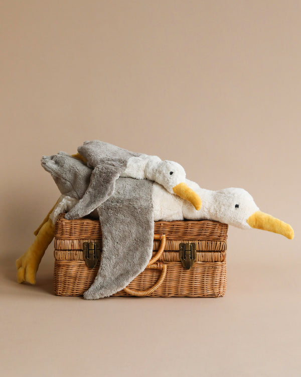 Two Senger Naturwelt Cuddly Animal - Seagulls made from organically grown cotton are lying on a wicker picnic basket against a plain beige background. The seagulls appear soft, with one draped over the edge of the basket.