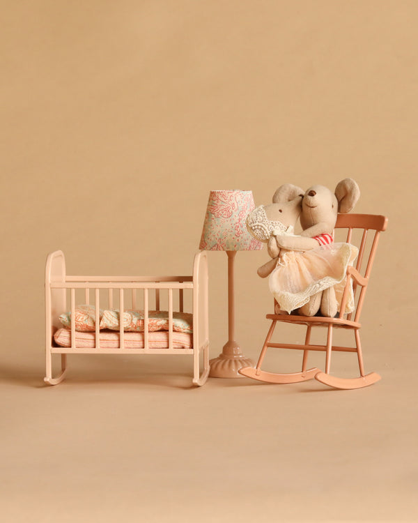 Two Maileg mice from the Maileg Big Sister and Me Nursery Set sit on a peach rocking chair next to a matching crib, lamp, and side table, all arranged against a beige background. The scene is styled like a cozy nursery, showcasing charming Maileg details throughout.