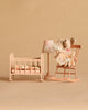 Two Maileg mice from the Maileg Big Sister and Me Nursery Set sit on a peach rocking chair next to a matching crib, lamp, and side table, all arranged against a beige background. The scene is styled like a cozy nursery, showcasing charming Maileg details throughout.