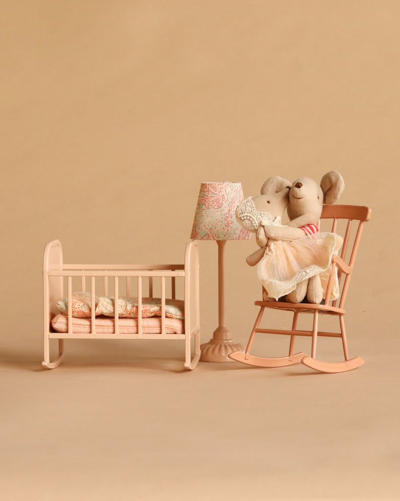 Two Maileg mice from the Maileg Big Sister and Me Nursery Set sit on a peach rocking chair next to a matching crib, lamp, and side table, all arranged against a beige background. The scene is styled like a cozy nursery, showcasing charming Maileg details throughout.
