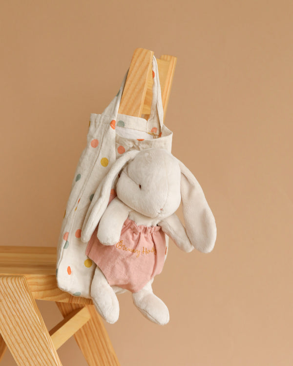 A Maileg Bunny Holly hanging from a wooden chair, clad in a pink shirt and inside a polka dot bag decorated with colorful polka dots, against a soft beige background.