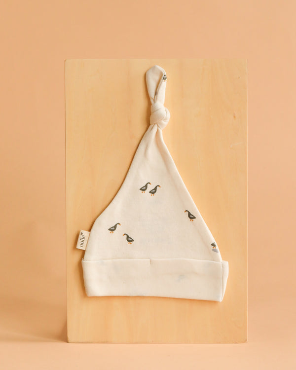 The Organic Baby Hat – Runner Ducks is a soft, cream-colored newborn hat made from GOTS-certified organic cotton with a top knot and playful duck prints, shown on a light wooden board against a beige background.