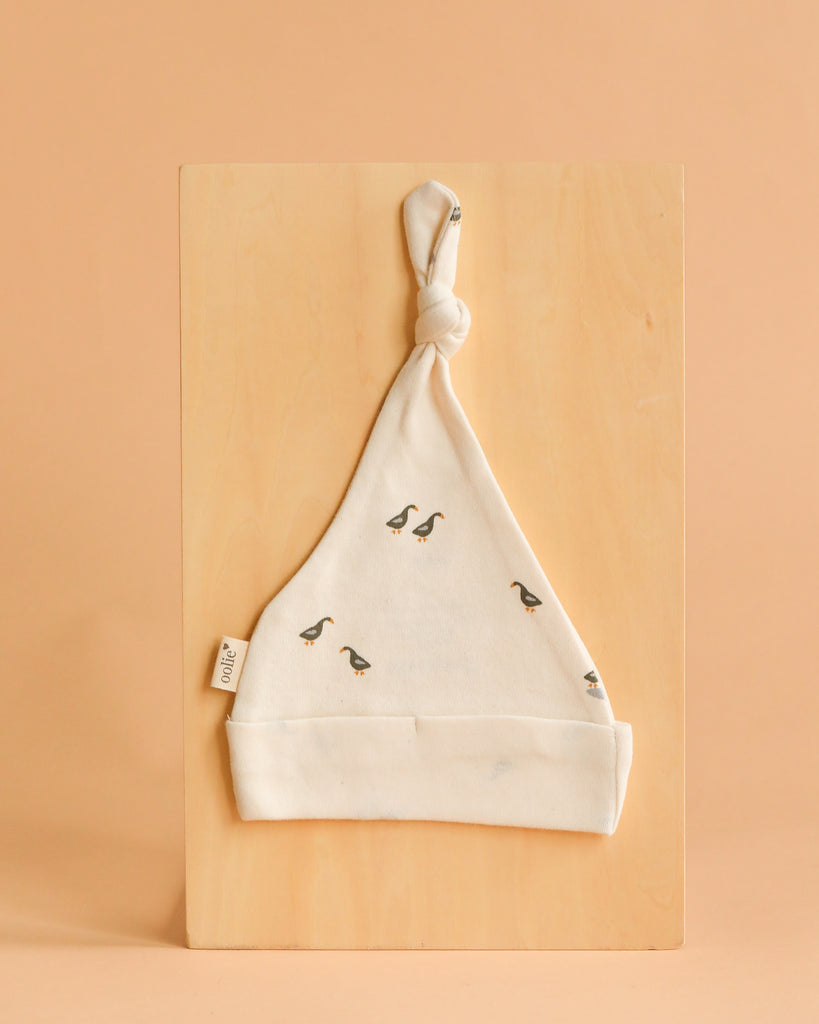 The Organic Baby Hat – Runner Ducks is a soft, cream-colored newborn hat made from GOTS-certified organic cotton with a top knot and playful duck prints, shown on a light wooden board against a beige background.