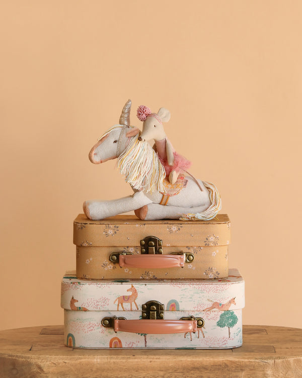 The Maileg Unicorn and Ballerina with Suitcase Set features a ballerina mouse on a unicorn, all displayed atop charming suitcases with animal and rainbow patterns, set against a beige background.