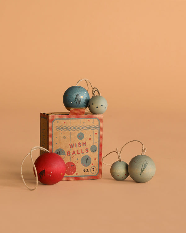 A Grapat Ornament Set - Wish Balls sits on a peach surface, surrounded by five colorful stringed ornaments—red, blue, and gray—arranged around and atop the box.