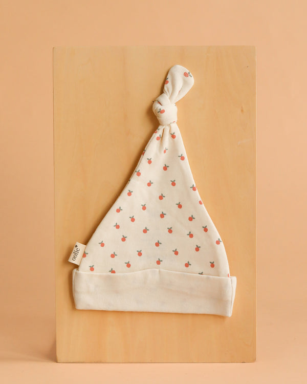 The Organic Baby Hat – Little Peach is a soft, white GOTS-certified organic cotton hat with a top knot and a cute red cherry print. It’s shown on a light wooden board with a beige background and has a small side fabric label.