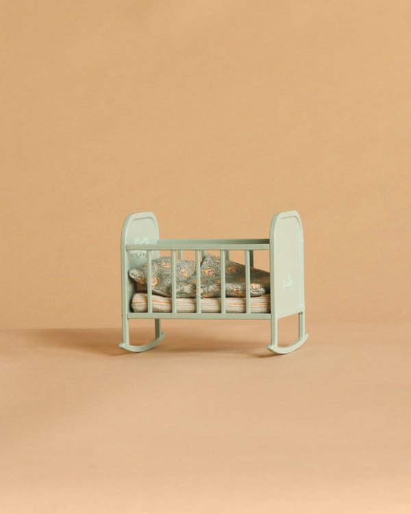 A Maileg Cradle, My in Blue Mint with a patterned pillow and blanket sits on a plain beige background.