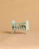 A Maileg Cradle, My in Blue Mint with a patterned pillow and blanket sits on a plain beige background.