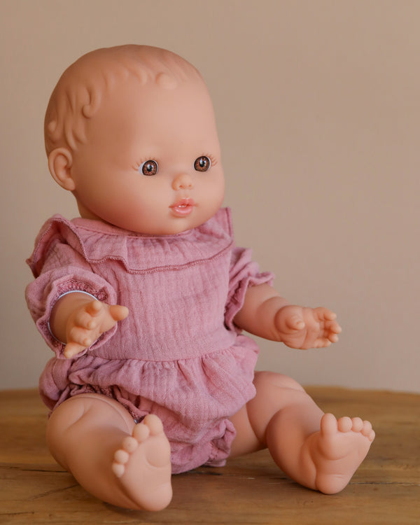 A Minikane Doll (13") - Faustine with short hair sits on wood. It’s made from phthalate-free vinyl and displayed in a pink romper with ruffled sleeves against a neutral background. The doll's open eyes, spread hands, and feet invite imaginative playtime. Clothing is sold separately.