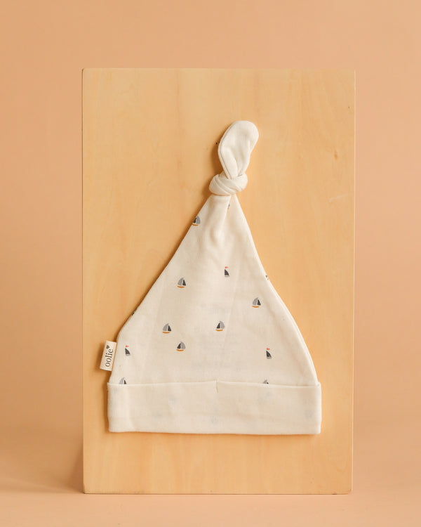 The Organic Baby Hat – Schooner is a soft, cream-colored GOTS certified organic cotton hat with tiny sailboat patterns and a knotted top, shown flat on a light wooden board against a peach background.