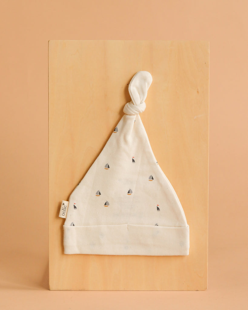 The Organic Baby Hat – Schooner is a soft, cream-colored GOTS certified organic cotton hat with tiny sailboat patterns and a knotted top, shown flat on a light wooden board against a peach background.