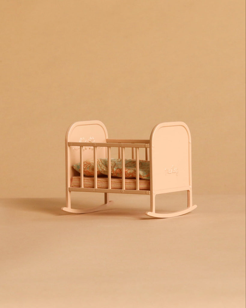 The Maileg Big Sister and Me Nursery Set, featuring a small, light pink wooden cradle with floral bedding, is displayed on a beige background.