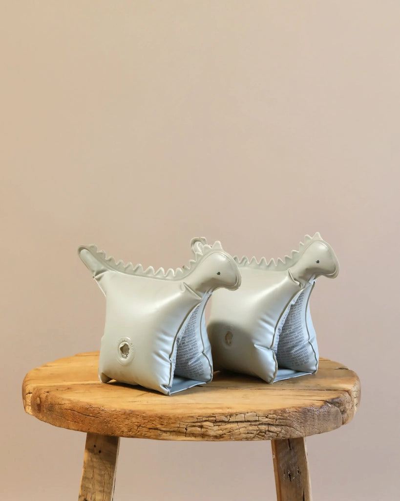A pair of Inflatable Water Wings - Dinosaur Shape for children aged 3-6 years, crafted from durable PVC and colored light gray, are placed on a round wooden stool against a light brown wall background. These floaties are designed to help children stay afloat in water.