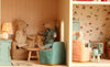 Three fabric mice lounge in the cozy living room and dining area of the Maileg Fully Furnished Farmhouse Set + Surprise Gift, complete with pastel Maileg Mouse Furniture, patterned wallpaper, a lamp, and a tiny television.